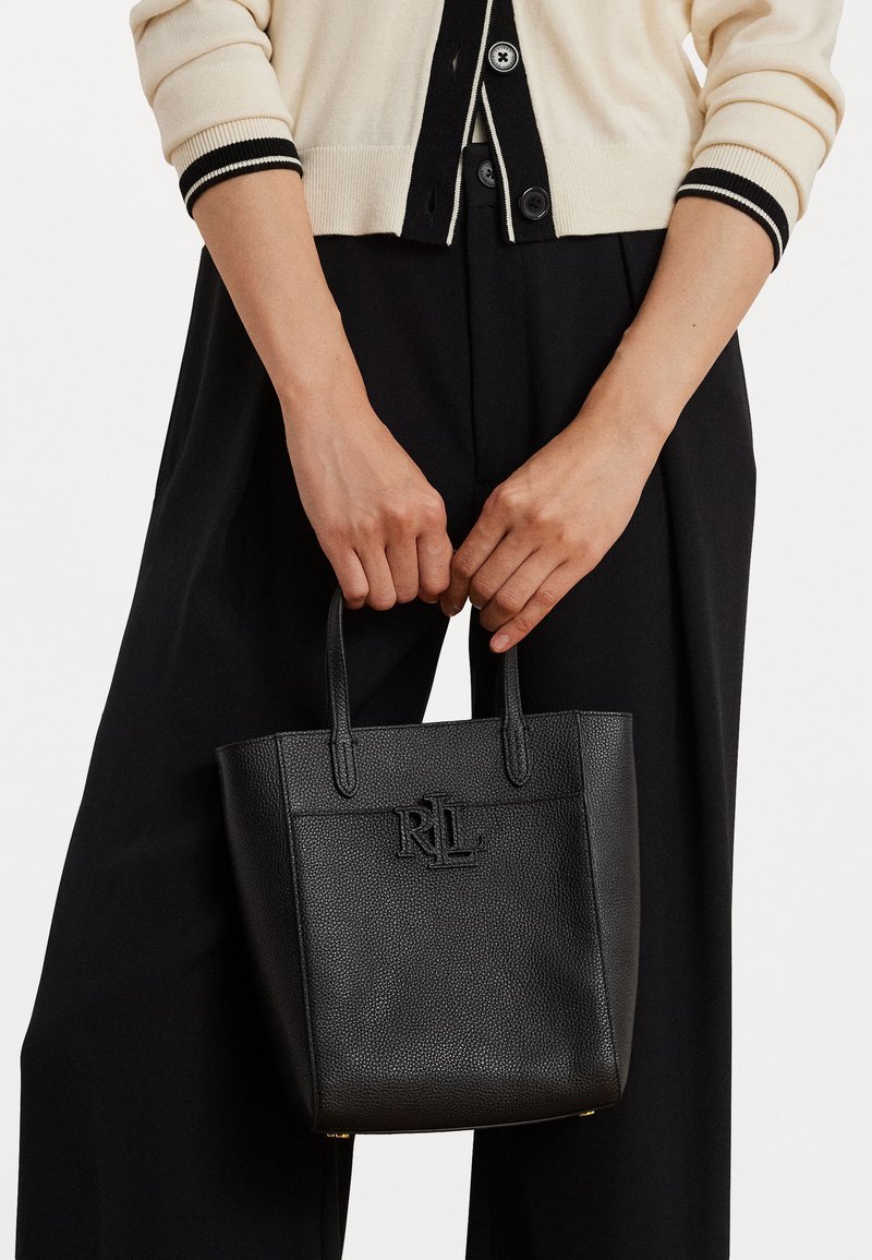 Melna teksturēta ādas rokassoma ar logo detaļu, kurai ir divas īsas rokas. To tur persona, kas ģērbusies bežā džemperī un melnās biksēs.