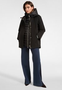 Zwarte waterdichte jas met een afneembare puffer binnenlaag, rits- en drukknoopsluitingen en subtiele gouden accenten, gecombineerd met een wijde denim spijkerbroek.