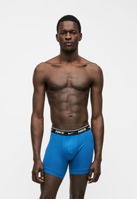 Αθλητικός σκουρόχρωμος άνδρας στέκεται, φοράει μπλε μποξεράκια Nike με μαύρη μέση σε απλό ανοιχτό φόντο.