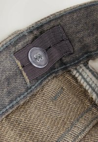 Rugalmas szövetből készült derékszalag, áttetsző műanyag gombbal, amely egy állítható gomblyuk szalaghoz csatlakozik, barna texturált nadrág belsejében.