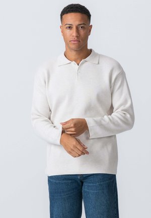Hvid langærmet polo-shirt med krave, to knapper og oprullede manchetter. Kombineret med blå denim jeans, vist foran en ensfarvet baggrund.