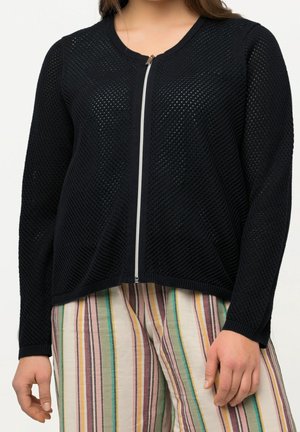 Kvinde iført en sort tekstureret cardigan med lynlås kombineret med højtaljede bukser med lodrette striber i pastelfarver.