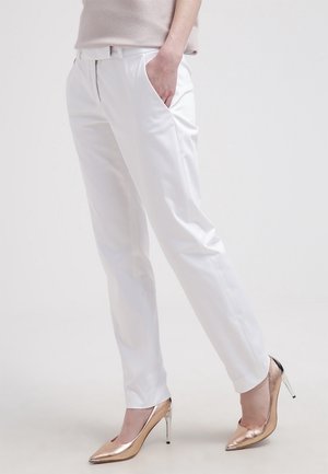 Vrouw draagt witte, op maat gemaakte broek met voorzakken en metallic rosegouden hoge hakken, staand tegen een effen achtergrond.