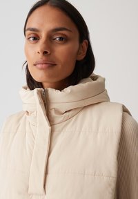 Beige gefütterte Weste mit hochgeschlossenem Kragen, Reißverschluss und dezenten Stickdetails; leichtes Material mit glatter Textur.
