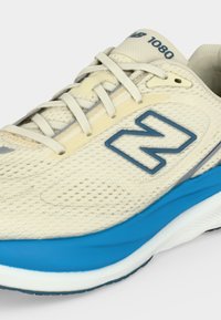 Ανοιχτό μπεζ αθλητικό παπούτσι New Balance 1080 με μπλε λογότυπο και ενδιάμεση σόλα, με επάνω μέρος από δίχτυ και λευκά κορδόνια.