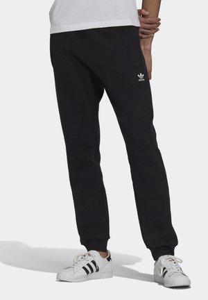 Person trägt schwarze Adidas-Sweatpants mit weißem Logo und weiße Adidas-Sneaker mit schwarzen Streifen, steht vor einem hellen Hintergrund.