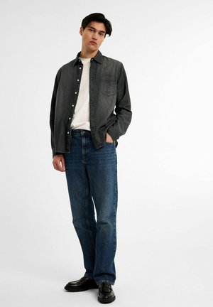 Dark grey button-up shirt over a white t-shirt, paired with blue denim jeans. Model stands with hands in pockets, wearing black shoes.