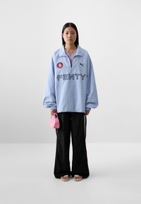 Model wearing an oversized light blue Fenty sweatshirt, black wide-leg pants, and pink handbag, standing against a minimalistic background.