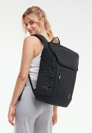 Nuori nainen, jolla on vaaleat hiukset, pukeutunut valkoiseen toppiin ja harmaisiin housuihin, kantaa suurta mustaa suorakaiteen muotoista reppua selässään.