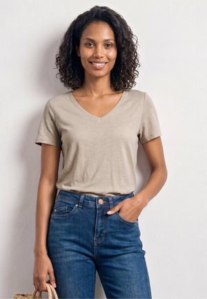 Uśmiechnięta kobieta z kręconymi włosami, ubrana w beżowy T-shirt z dekoltem w szpic i niebieskie dżinsy, stojąca na jasnym tle, trzymająca plecioną torbę.