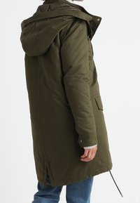 Parka à capuche vert olive avec fermeture éclair, poches latérales et léger effet queue-de-pie. Tissu épais avec une finition lisse et une doublure matelassée.