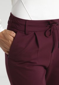 Burgundské kalhoty mají elastický pás se šňůrkou, boční kapsy a hladký, textura. U pásu jsou jemné sklady.