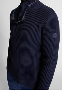 Pull en mailles côtelées bleu marine avec un col à demi-zip, un patch logo sur la manche et des lunettes de soleil accrochées au décolleté. Matière en tricot texturé.