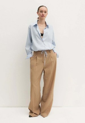 Donna che indossa una camicia a righe azzurro chiaro sopra un reggiseno nero e pantaloni larghi beige larghi, in piedi contro un muro bianco semplice.