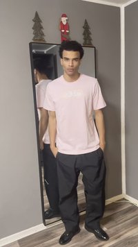 Rozā t-krekls ar priekšpuses grafiku, melnas brīvā stila bikses un spīdīgas melnas kurpes. Spogulis atspoguļo pilnu apģērbu un apkārtējo dekoru.