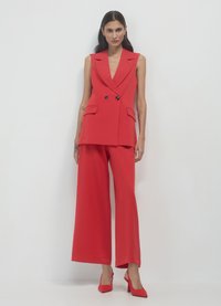 Roter, maßgeschneiderter ärmelloser Blazer mit doppelten Knöpfen und Taschen, kombiniert mit weit geschnittenen Hosen in passender Farbe. Dazu Stilettos.