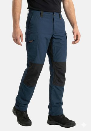 Mężczyzna w granatowych spodniach roboczych z czarnymi wzmocnionymi łatami na kolanach i czarnych butach trekkingowych, stojący na białym tle.