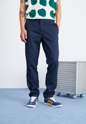 Navy blue trousers, tailored fit, cotton blend. Paired with blue sneakers featuring yellow stripes and white laces. Lightweight, modern design.