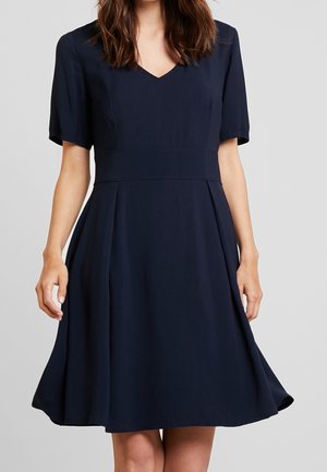 Vrouw draagt een knielange marineblauwe jurk met korte mouwen en V-hals, staat tegen een effen lichtgrijze achtergrond.