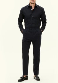 Camicia blu navy con bottoni abbinata a pantaloni su misura, entrambi realizzati in tessuto liscio. Le scarpe sono mocassini neri con dettagli a nappina.