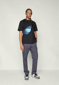 Sling bag in iridescent blue material, worn over a black cotton t-shirt. Model in grey trousers and black and white sneakers.