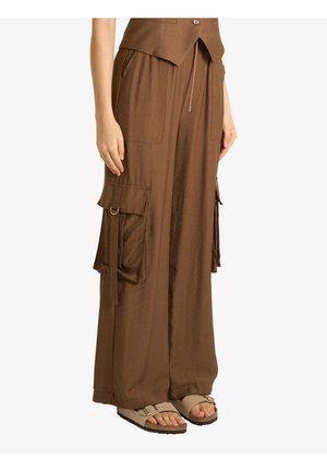 Persona che indossa pantaloni cargo marroni a gamba larga con grandi tasche e sandali beige aperti in punta, in piedi su sfondo bianco.
