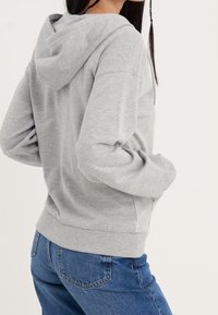 Sudadera gris con capucha, de textura suave, con mangas largas y bolsillo tipo canguro. Combinada con jeans azules de cintura alta.