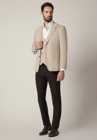 Mężczyzna w beżowym żakiecie i kamizelce, białej koszuli, ciemnych spodniach i brązowych butach eleganckich, stojący na jednolitym jasnym tle.