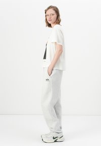 Vrouw die zijwaarts staat in een wit T-shirt met opdruk, lichtgrijze joggingbroek met logo en witte sneakers tegen een effen witte achtergrond.