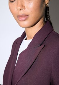 Bordeauxroter Blazer mit einem eleganten Design, ausgestattet mit einem spitzen Kragen und strukturiertem Stoff. Getragen über einem weißen Oberteil, das eine minimalistische Ästhetik zeigt.