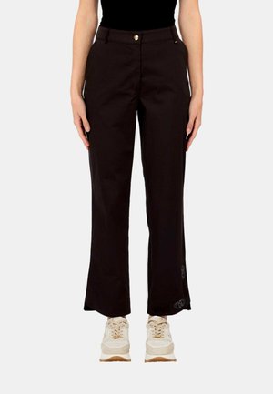 Donna che indossa pantaloni neri a gamba dritta con bottone dorato e sneakers beige, in piedi davanti a uno sfondo bianco semplice.