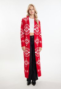 Roter gestrickter Cardigan mit weißen geometrischen Mustern, langen Ärmeln, offener Front und lockerer Passform, kombiniert mit schwarzen Hosen und Absatzschuhen.