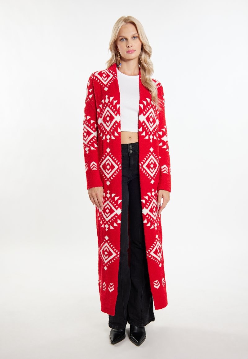 Roter gestrickter Cardigan mit weißen geometrischen Mustern, langen Ärmeln, offener Front und lockerer Passform, kombiniert mit schwarzen Hosen und Absatzschuhen.