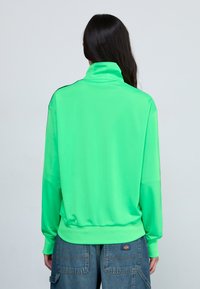 Neon grön sweatshirt med hög krage, långa ärmar och ribbad nederkant; har svarta axelinslag. Slät textur, avslappnad design.