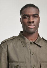 Camisa de botón verde oliva con cuello estructurado, dos bolsillos en el pecho y epauletas en los hombros. Tejido suave con un brillo sutil.