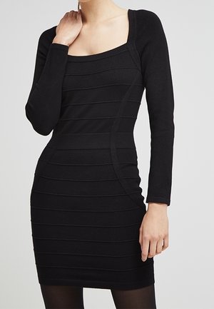 Frau trägt ein figurbetontes, langärmeliges schwarzes Kleid mit horizontalen, strukturierten Streifen und quadratischem Ausschnitt.