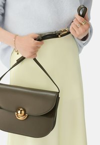 Tmavě zelená kožená kabelka s kulatým klopem a zlatými kovovými detaily. Popruh je nastavitelný a kabelku drží ruka blízko světlé sukně.