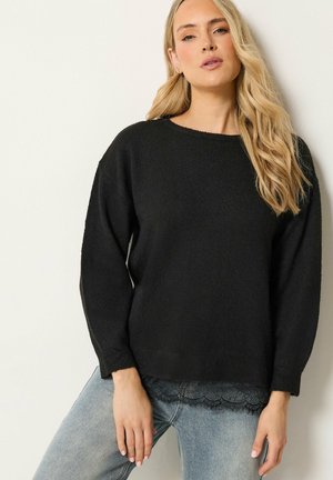 Vrouw met lang blond haar die een zwarte trui draagt over een top met kanten rand en lichtblauwe spijkerbroek tegen een effen achtergrond.