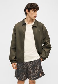 Jonge man met een olijfgroene jas, wit shirt en zwarte Nike-short met patroon, hand in zak, kijkend naar de zijkant.