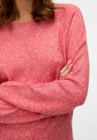 Pull en maille rose avec un mélange de textures. Il présente un col rond et de longues manches, les bras croisés, mettant en valeur le tissu doux.