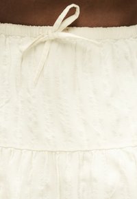 Jupe en tissu blanc avec taille élastique et nœud noué en haut, avec des motifs texturés verticaux subtils.