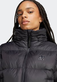 Giubbotto piumino nero con colletto alto, texture trapuntata orizzontale, chiusura con zip e piccolo logo riflettente sul petto. Tessuto morbido.