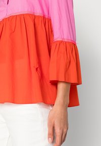 Top con blocchi di colore, con una sezione superiore rosa e una sezione inferiore arancione. Presenta maniche a balze e una vestibilità ampia. Il materiale appare leggero.