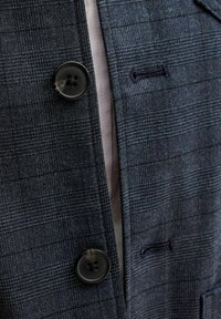 Tamsiai mėlynas languotas švarkas, pagamintas iš tekstūruotos medžiagos, su dviem dideliais juodais mygtukais ir subtilia atvira siūlės detalė prie apykaklės.