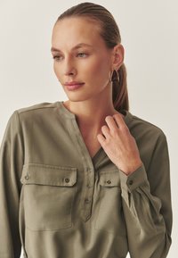 Olivgrüne Bluse mit Kragen, zwei Brusttaschen, Knopfleiste, langen Ärmeln und gerafften Schulterdetails. Weicher, glatter Stoff.