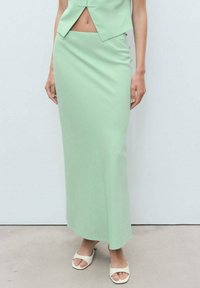Jupe maxi ajustée de couleur vert menthe en tissu lisse. Présente une fente sur le côté et une silhouette épurée, portée avec des sandales blanches ouvertes.