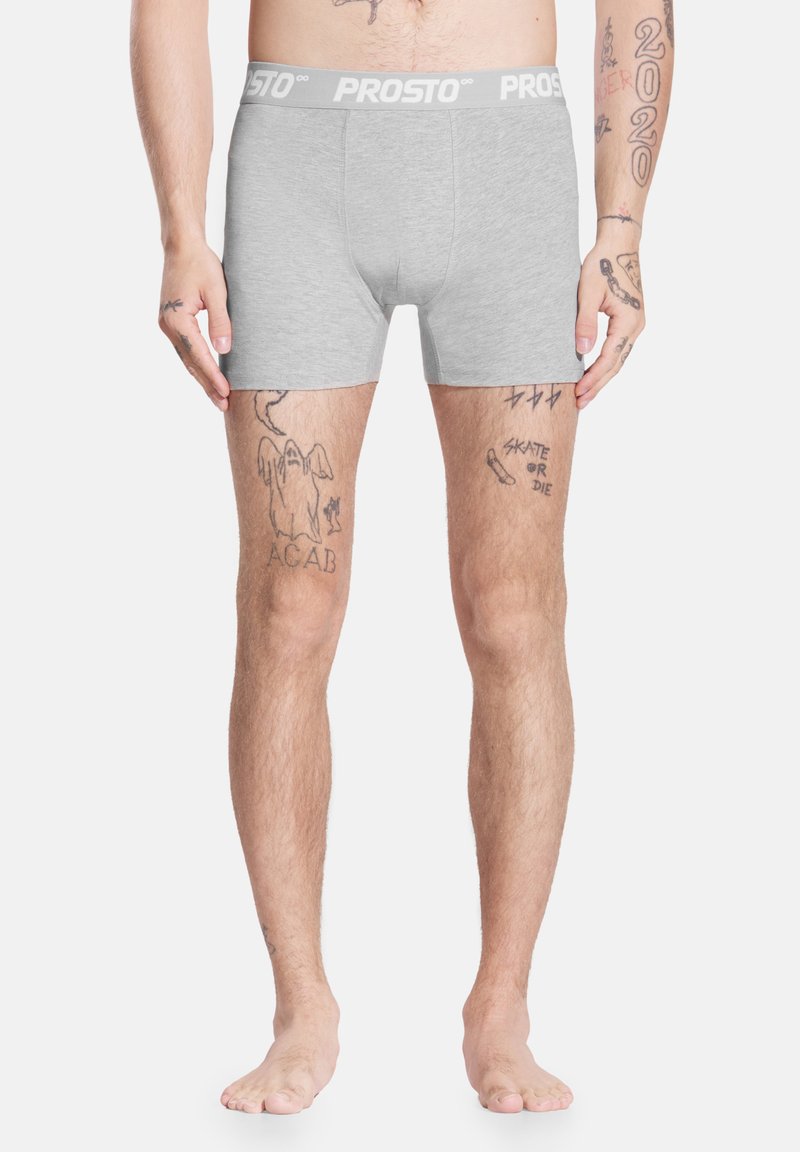 Szare bokserki z białym paskiem w talii z napisem "PROSTO", wykonane z gładkiego materiału, o dopasowanym kroju. Widoczne tatuaże na nogach.
