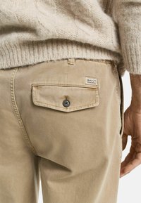 Pantalones de algodón beige con un bolsillo trasero que cuenta con un cierre de botón y una etiqueta que muestra la marca "GANT". Suéter de punto suave arriba.
