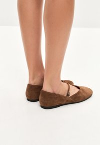 Zapatos planos de ante marrón con una correa en el tobillo, diseño de punta afilada y suela negra baja. Textura suave con un contorno simple.