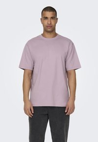 Camiseta de manga corta en lavanda claro, fabricada en suave algodón, con cuello redondo y un corte relajado, combinada con pantalones gris oscuro.